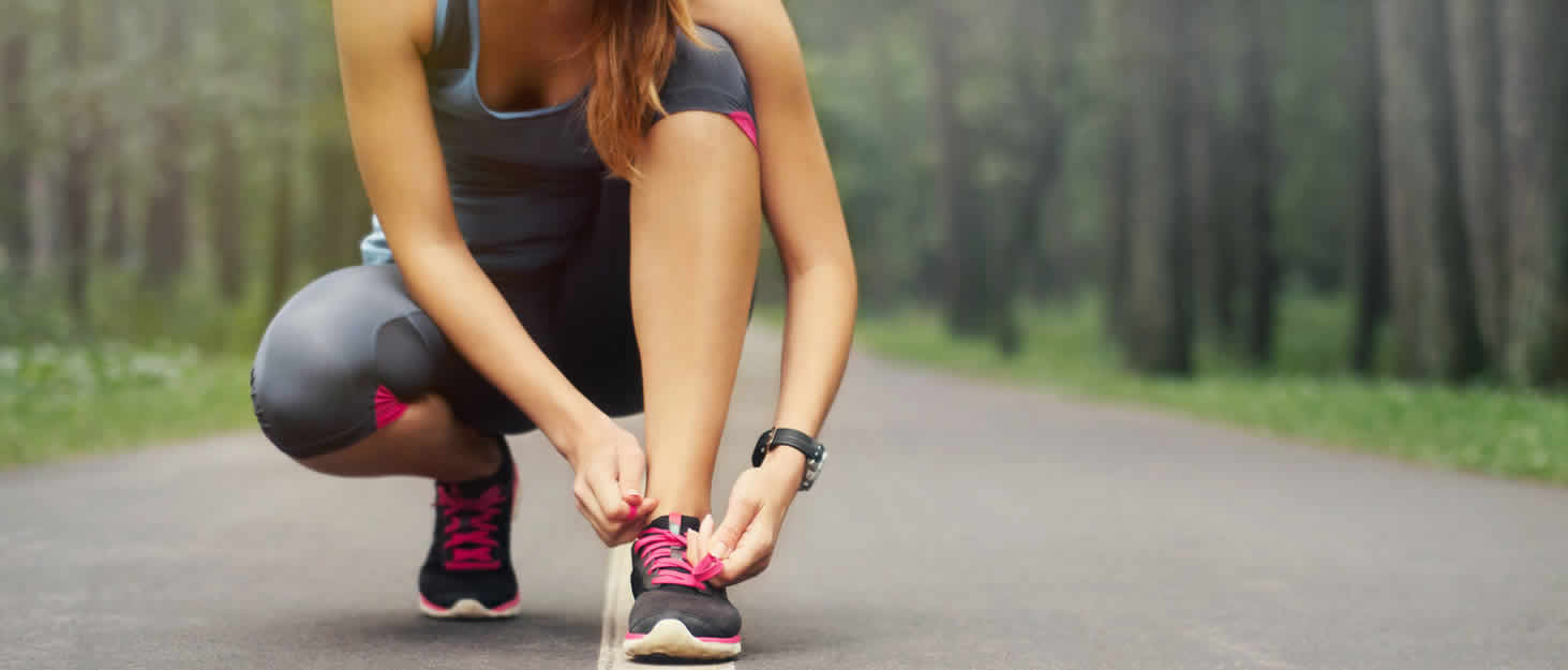 A woman kneeling down to tie her running shoe on a running path through the forest.